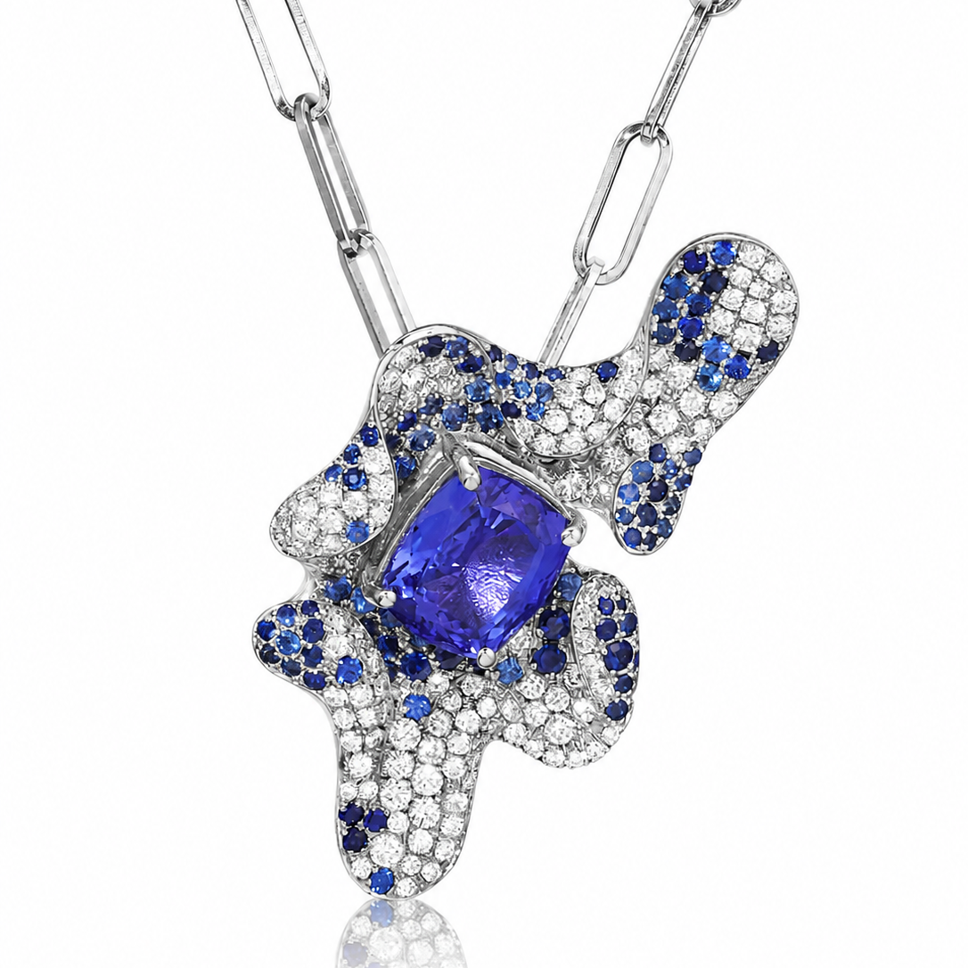 An abstract pendant necklace featuring a large central blue tanzanite gemstone surrounded by pavé-set blue sapphires and clear stones on a silver-toned chain, shown against a white background.