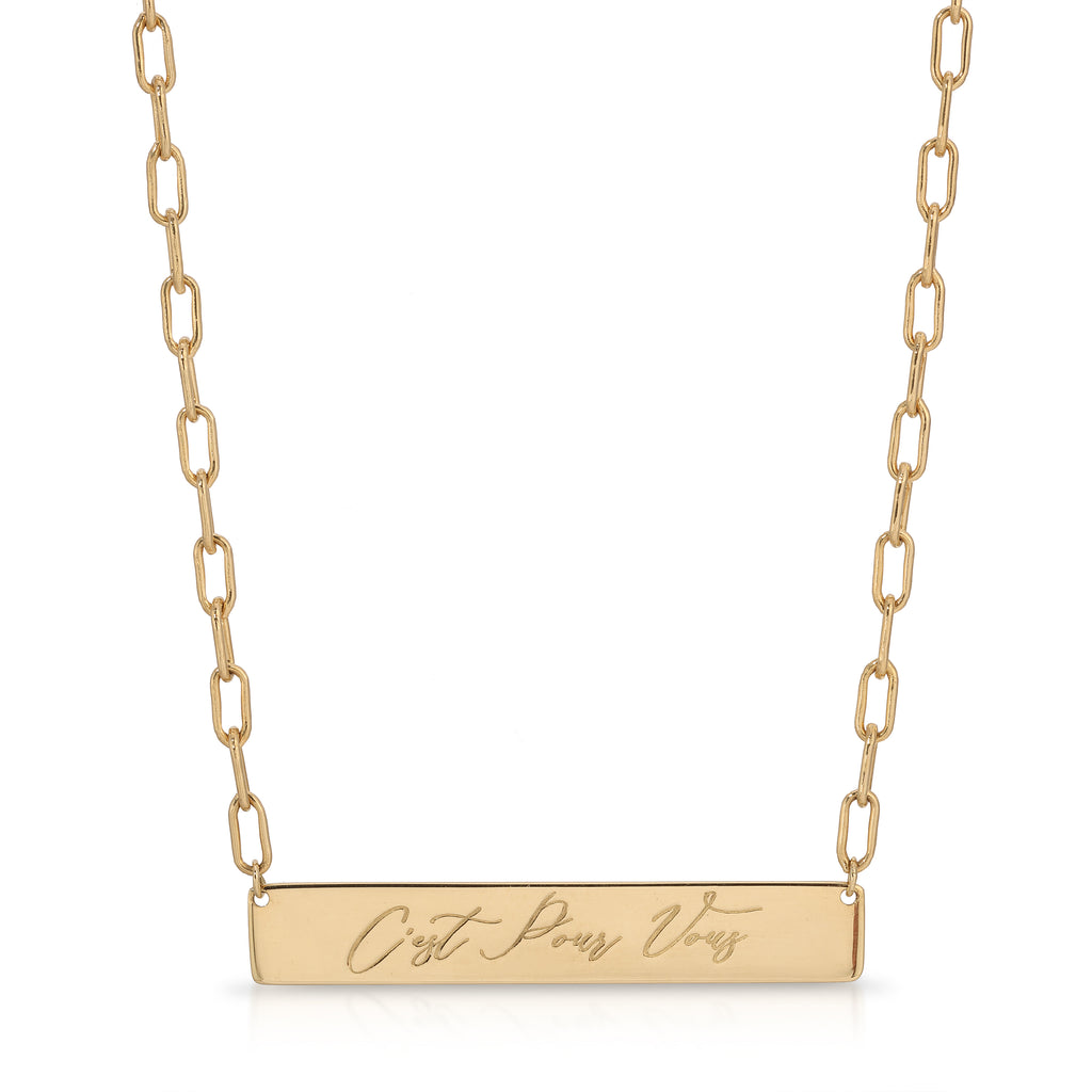 A gold bar necklace with a rectangular pendant engraved with the French phrase 'C'est Pour Vous', suspended from a gold paperclip chain on a white background.