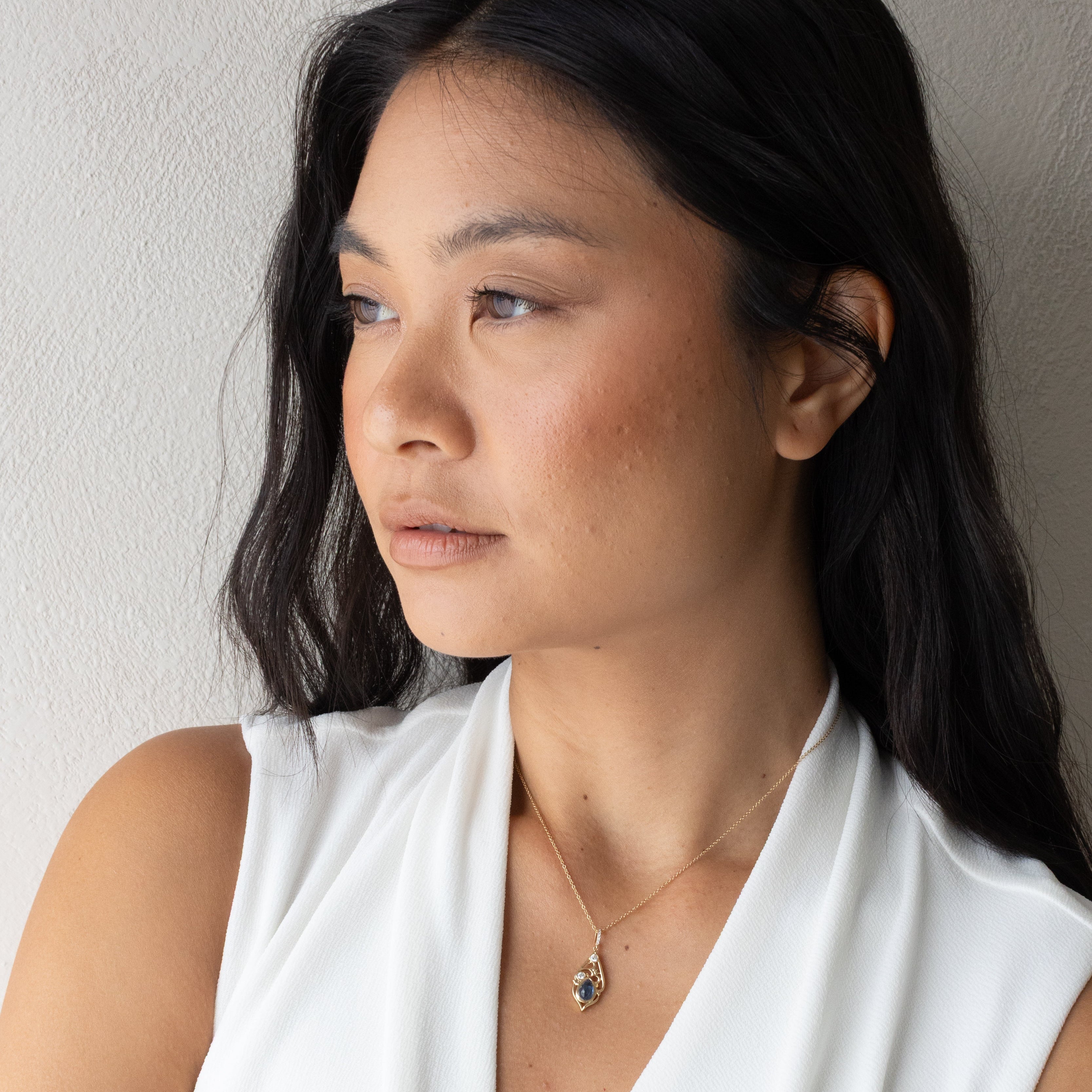 A woman with long black hair wearing a white sleeveless draped top and a delicate silver necklace, shown in a close-up portrait against a light textured wall.