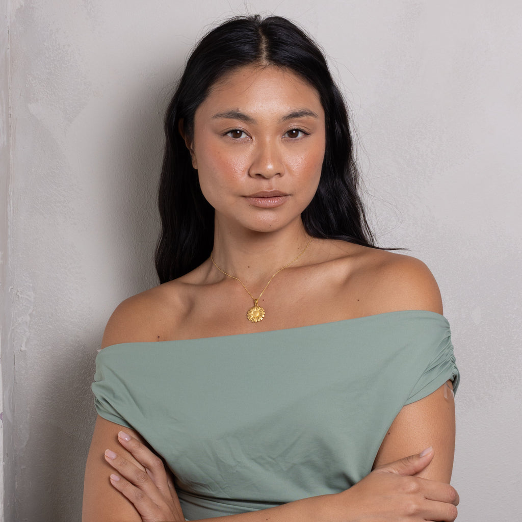 A woman with long dark hair wearing an off-the-shoulder sage green top and a gold pendant necklace, standing with her arms crossed against a textured light gray wall.