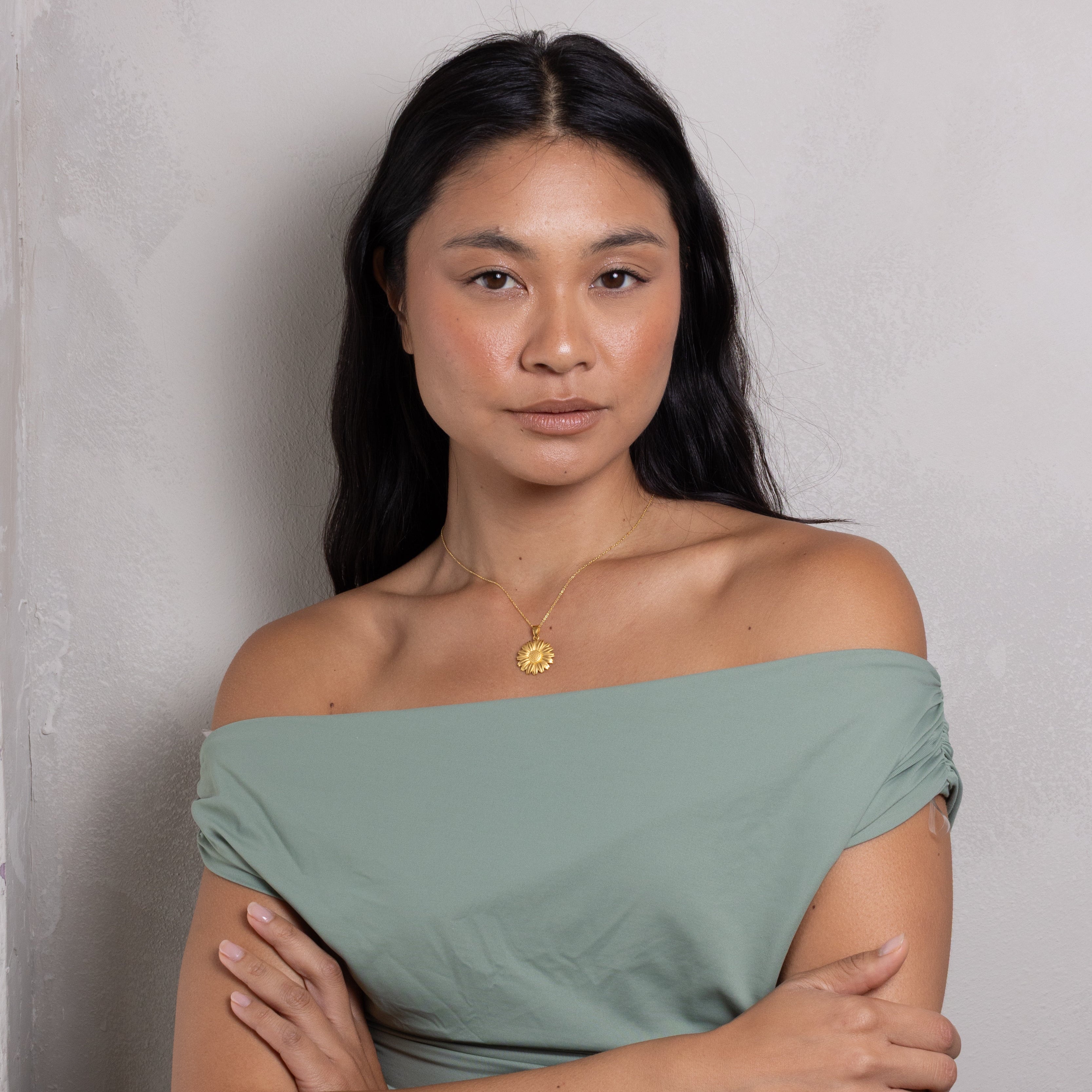 A woman with long dark hair wearing an off-the-shoulder sage green top and a gold pendant necklace, standing with her arms crossed against a textured light gray wall.