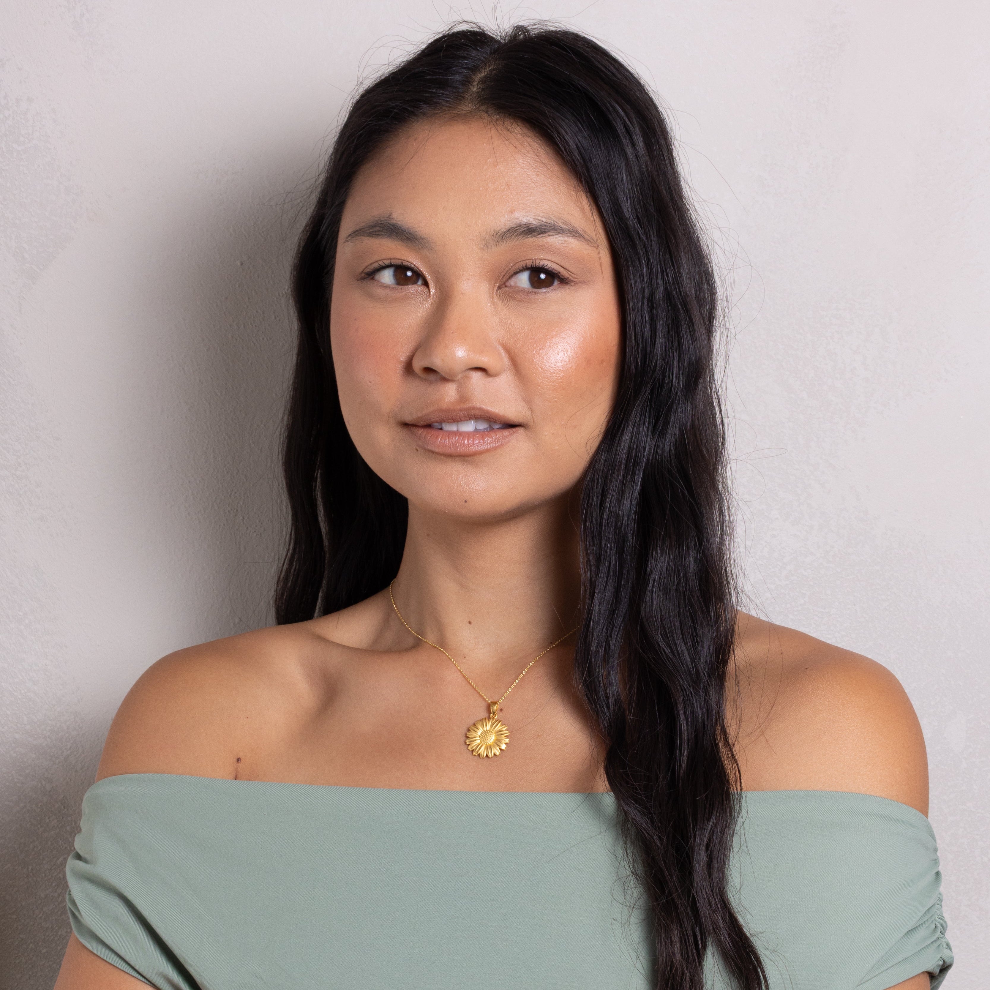 A woman with long dark hair wearing a sage green off-the-shoulder top and a gold sun pendant necklace, photographed against a neutral light gray wall.