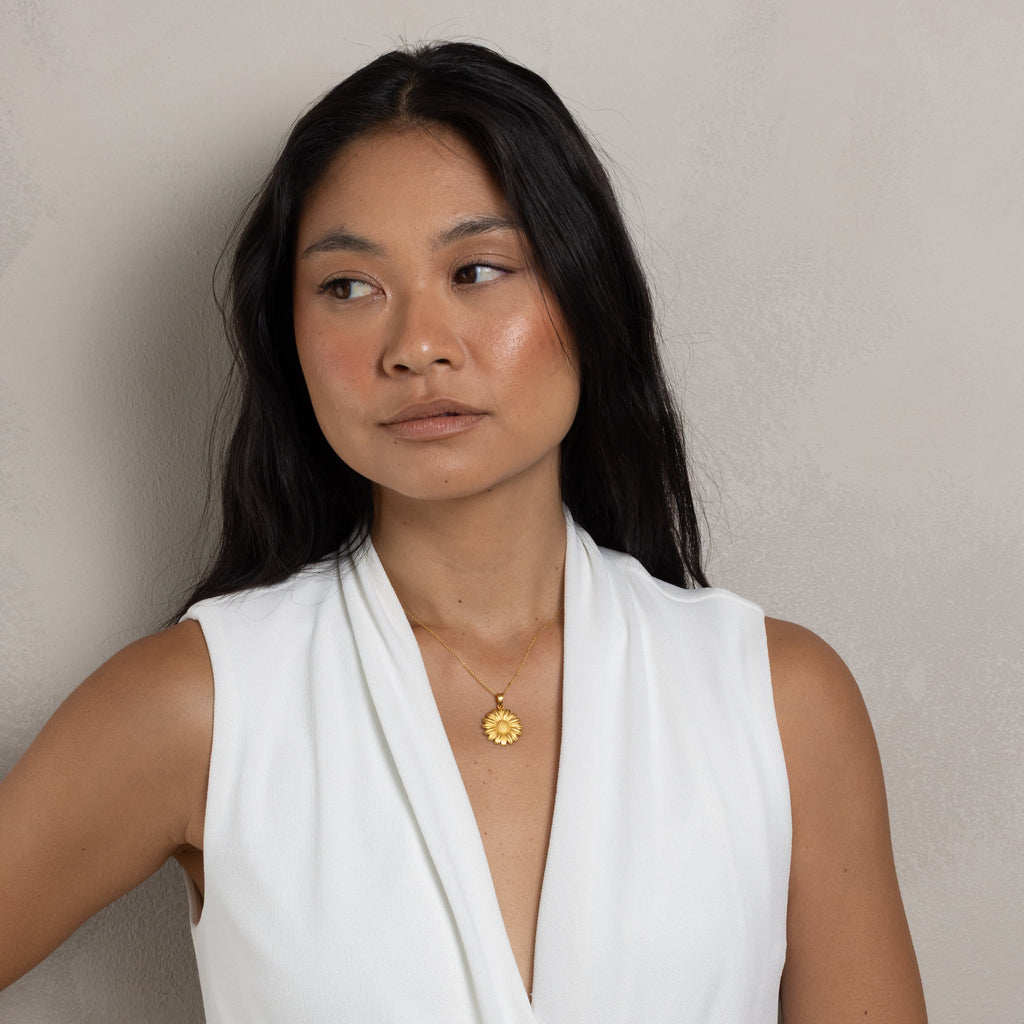 A woman with long black hair wearing a white sleeveless top and a delicate gold pendant necklace, posing against a neutral textured beige wall.