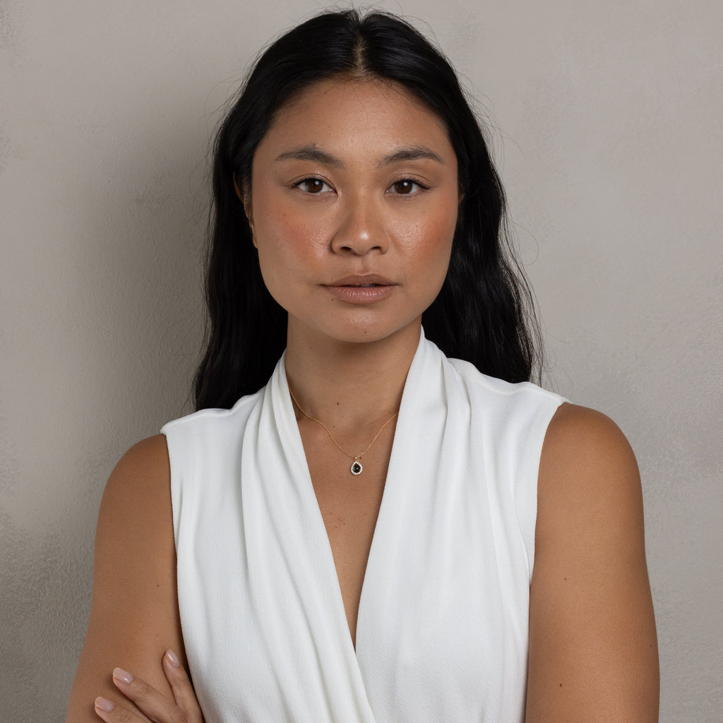 A woman with long black hair wearing a white sleeveless draped top, standing with her arms crossed against a plain light gray wall.