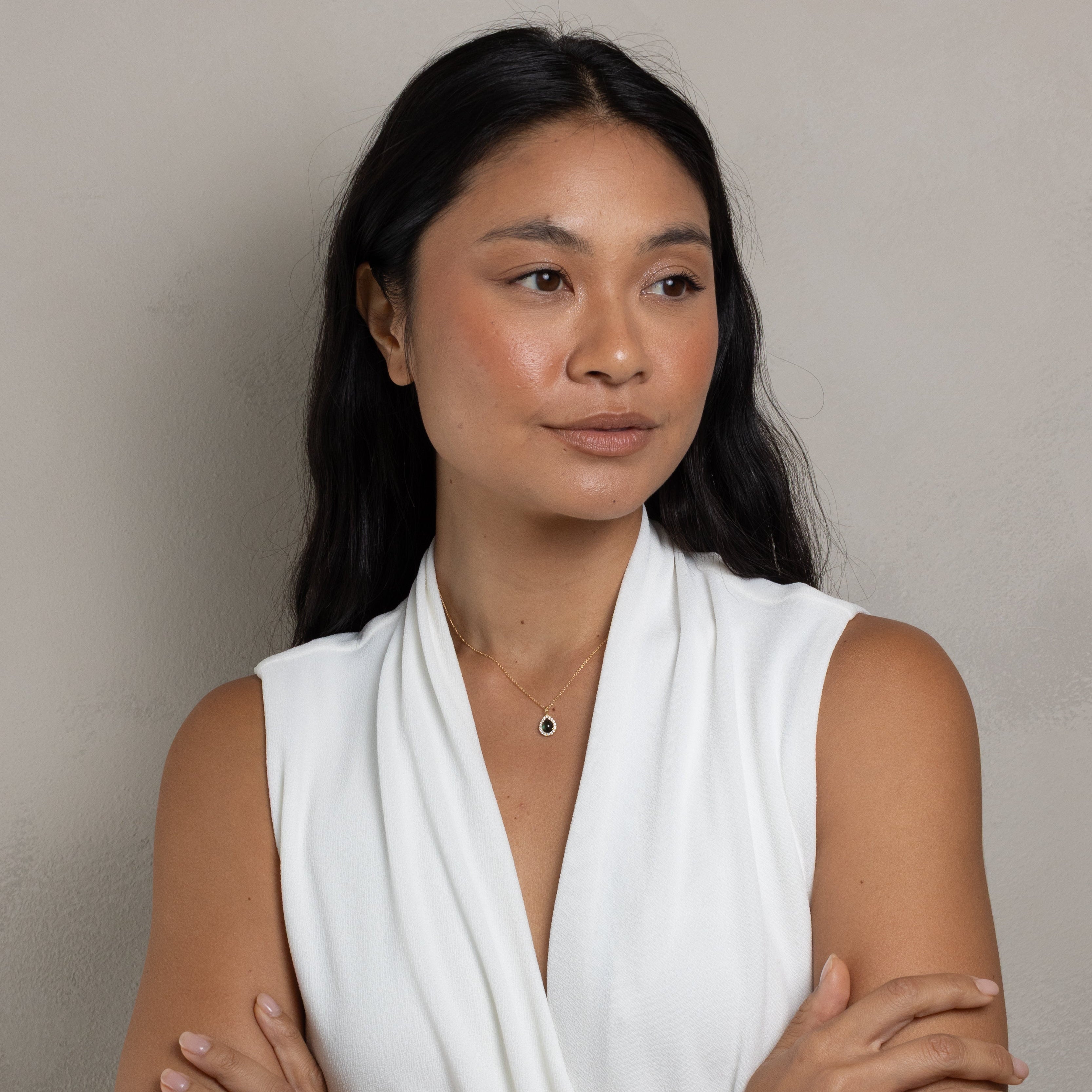 A woman with long dark hair wearing a white sleeveless draped top, standing with her arms crossed against a plain light gray wall.