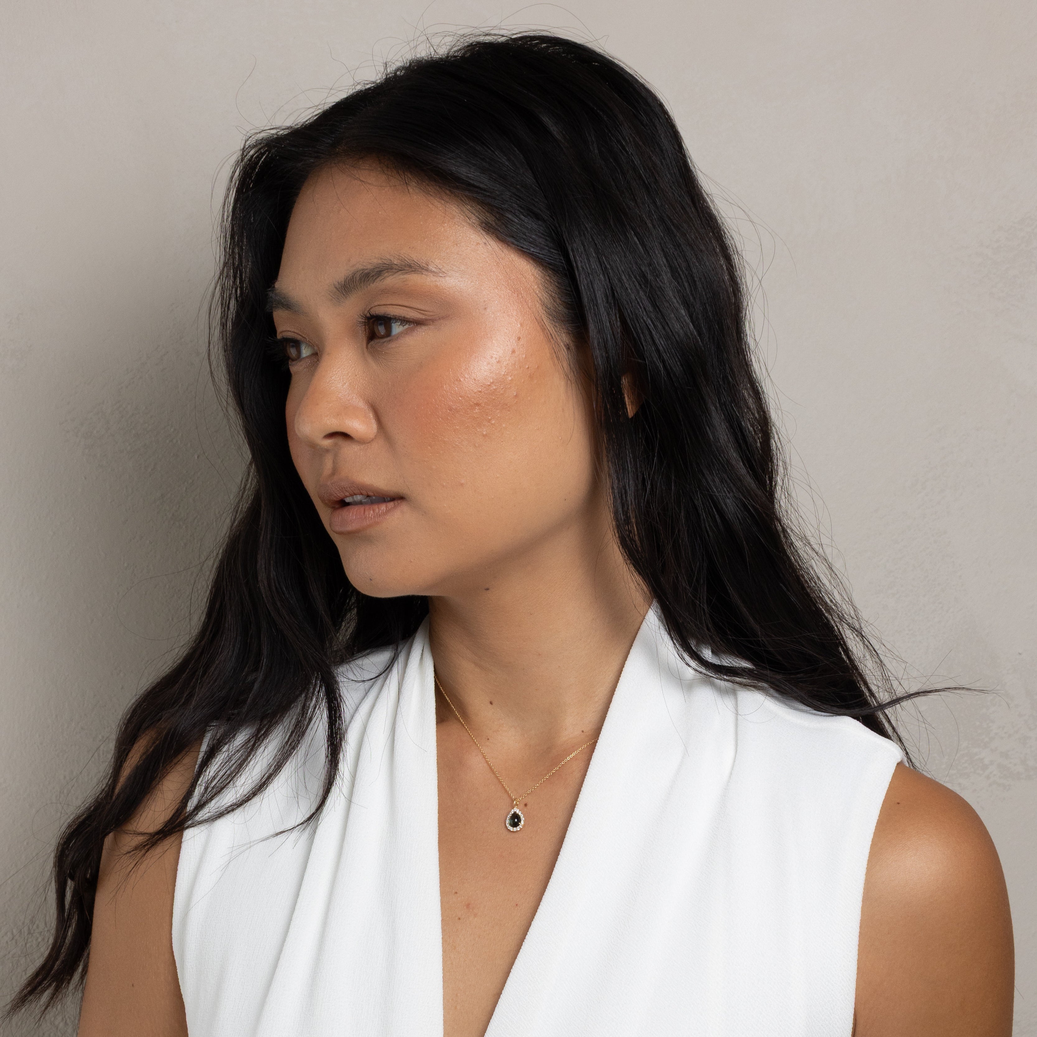 A woman with long dark hair wearing a white sleeveless wrap-style top, photographed against a plain light gray wall.