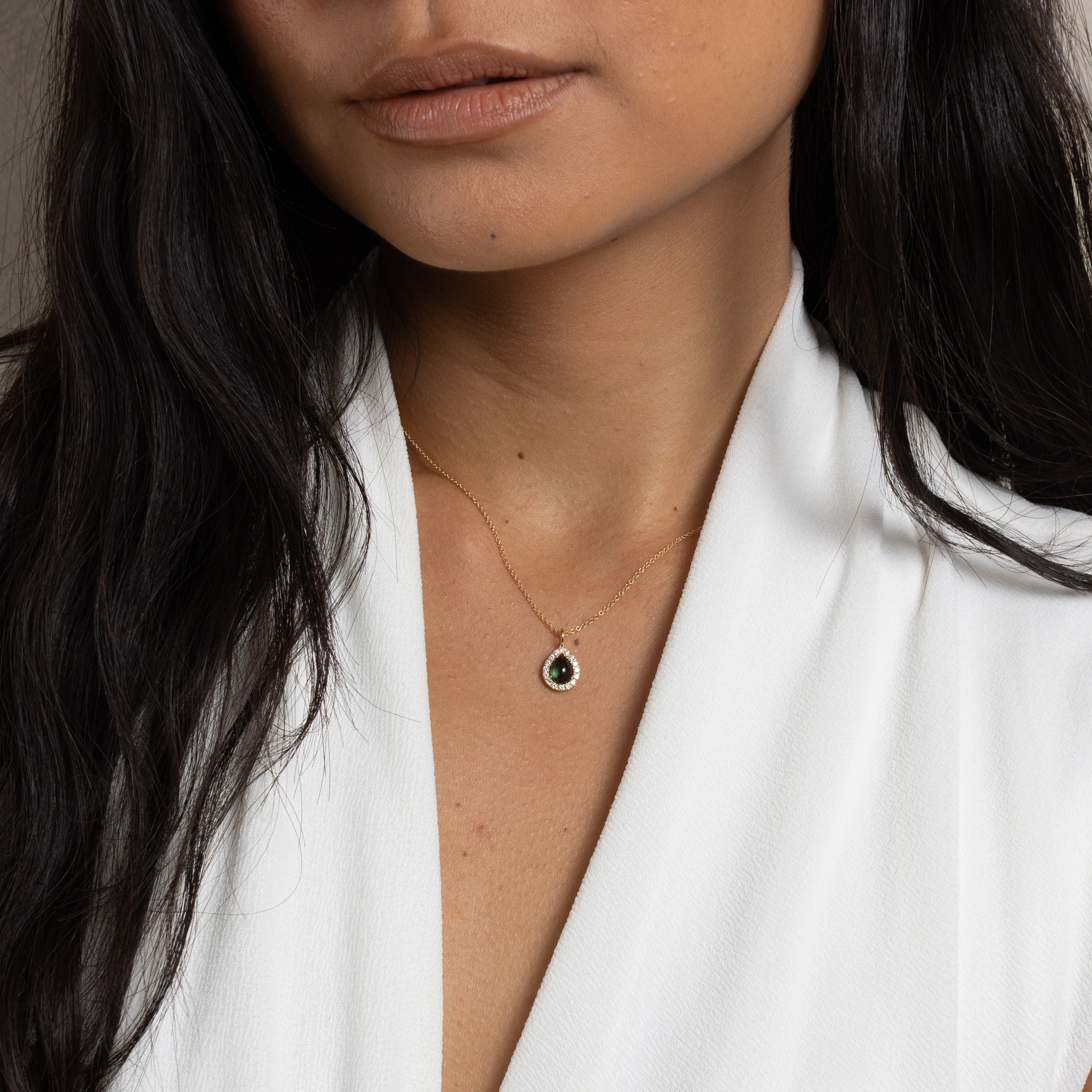 A close-up of a woman wearing a delicate gold necklace with a small teardrop-shaped pendant featuring a dark center, paired with a white sleeveless top.