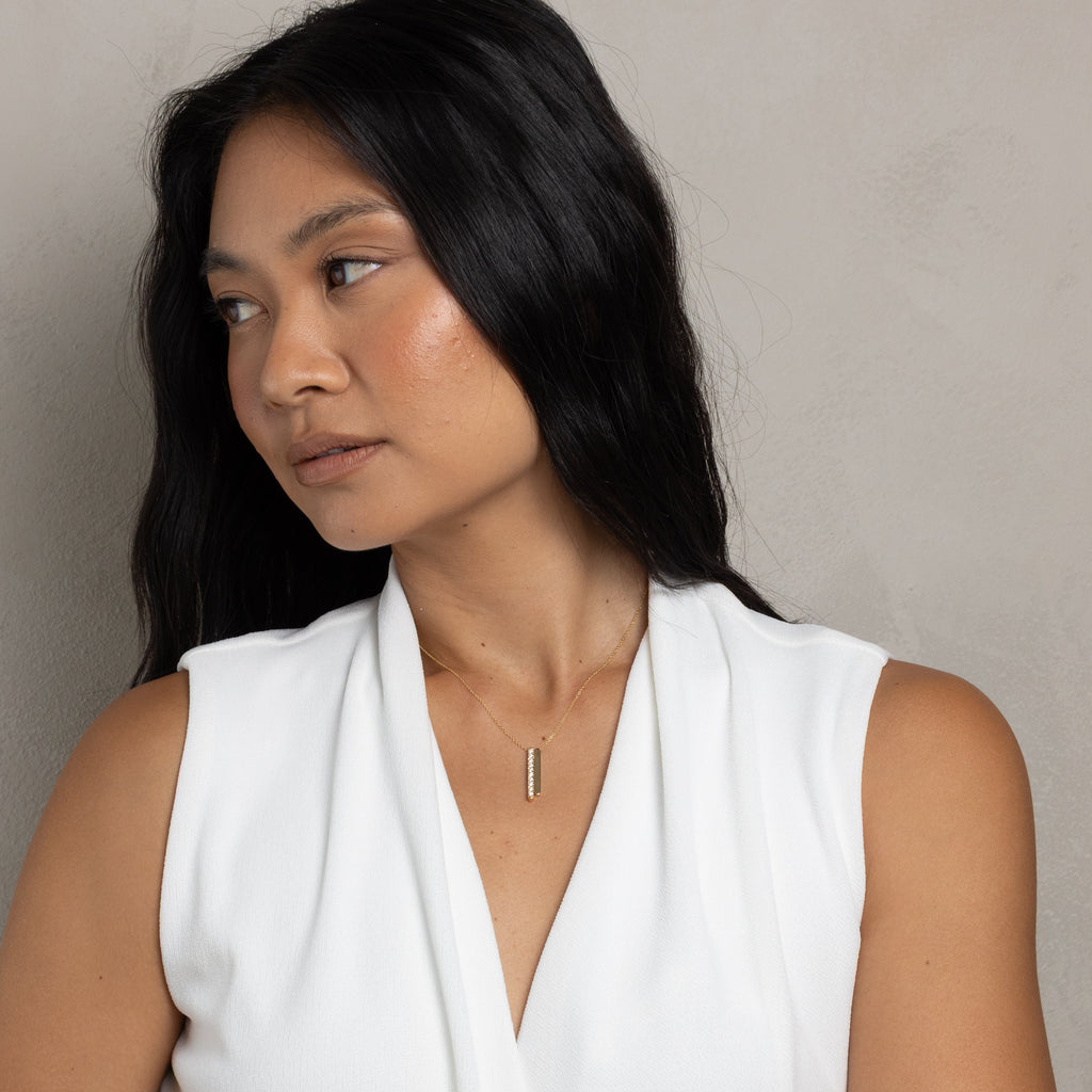 A woman with long black hair wearing a white sleeveless draped top and a thin vertical gold pendant necklace, photographed against a plain light gray wall.
