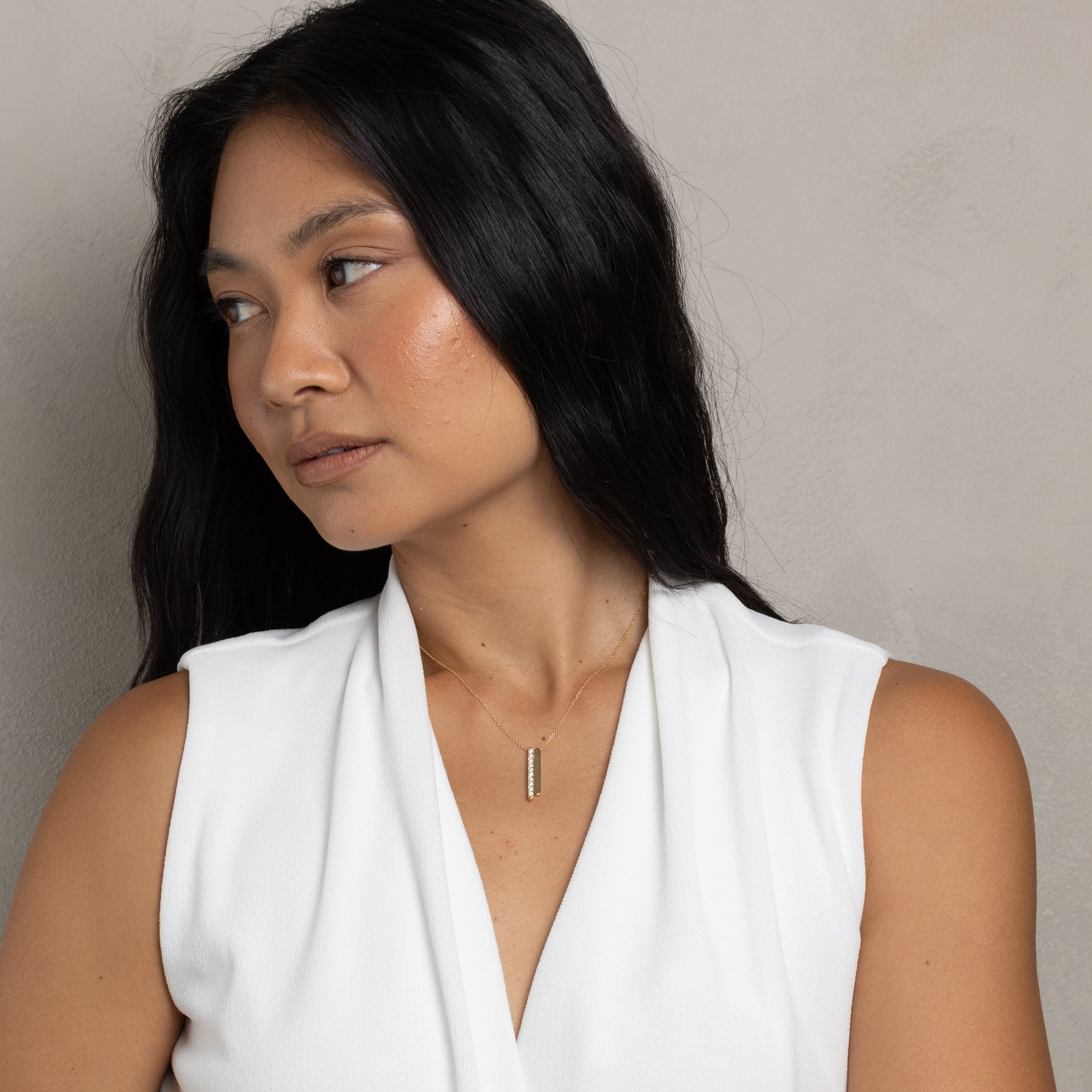 A woman with long black hair wearing a white sleeveless draped top and a thin vertical gold pendant necklace, photographed against a plain light gray wall.