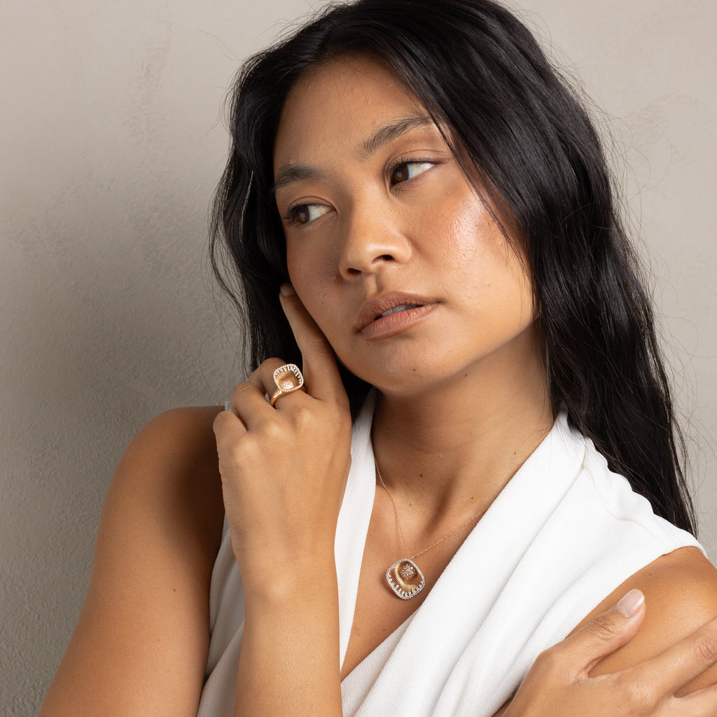A woman with long dark hair wearing a white draped top, showcasing a delicate gold necklace with a textured circular pendant and a matching ring on her finger, set against a neutral beige background.