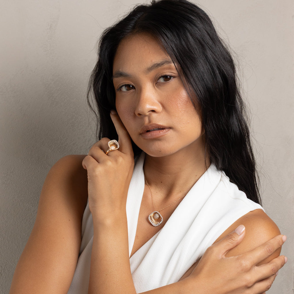 A woman with long black hair wearing a white draped top, a gold circular pendant necklace, and a gold ring, posing with her arms crossed against a neutral gray background.