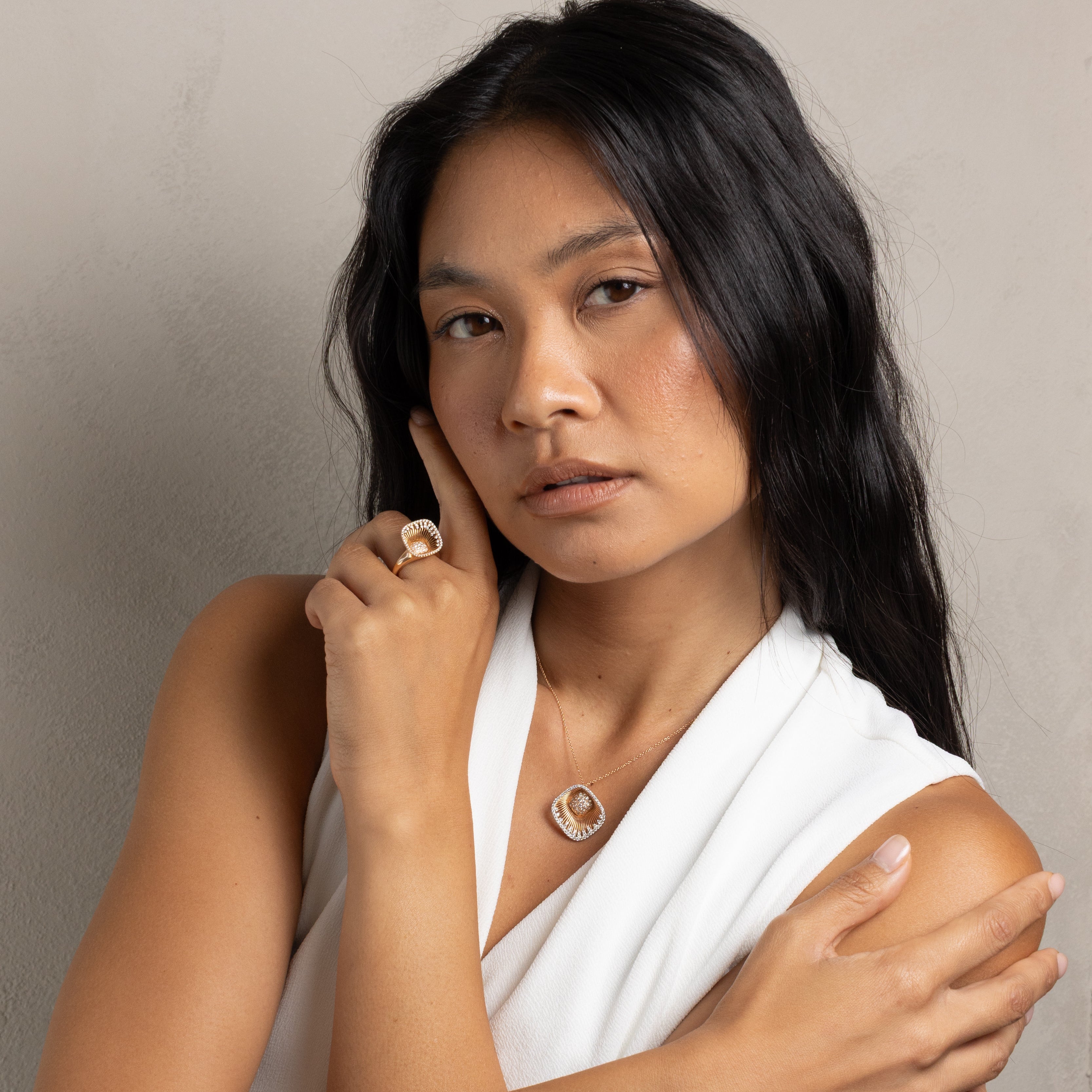 A woman with long black hair wearing a white draped top, a gold circular pendant necklace, and a gold ring, posing with her arms crossed against a neutral gray background.