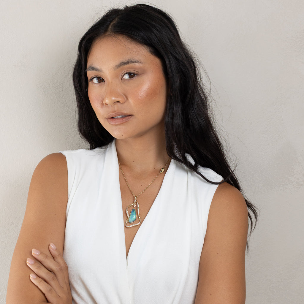A woman with long dark hair wearing a white sleeveless top and a gold pendant necklace with a turquoise-colored stone, posing with her arms crossed against a neutral light gray wall.