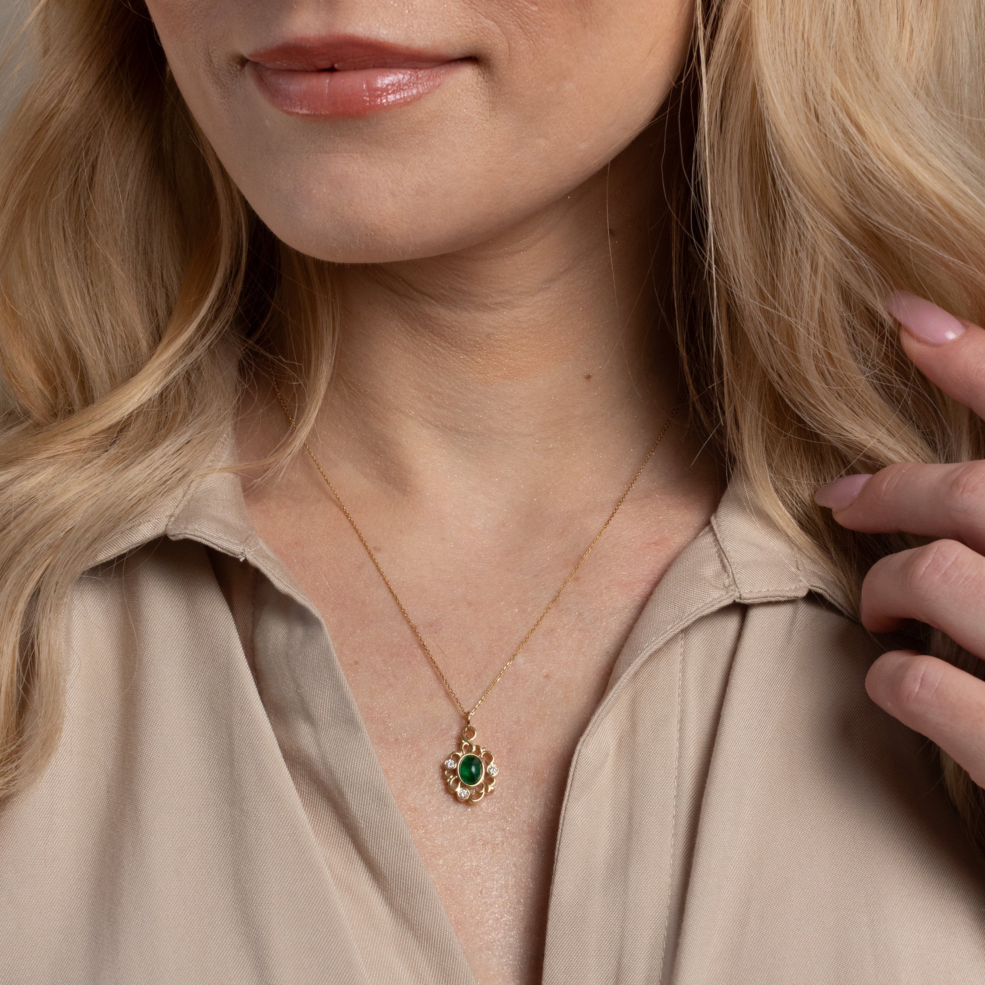 A close-up of a woman wearing a delicate gold necklace with a green heart-shaped pendant, paired with a beige collared blouse.