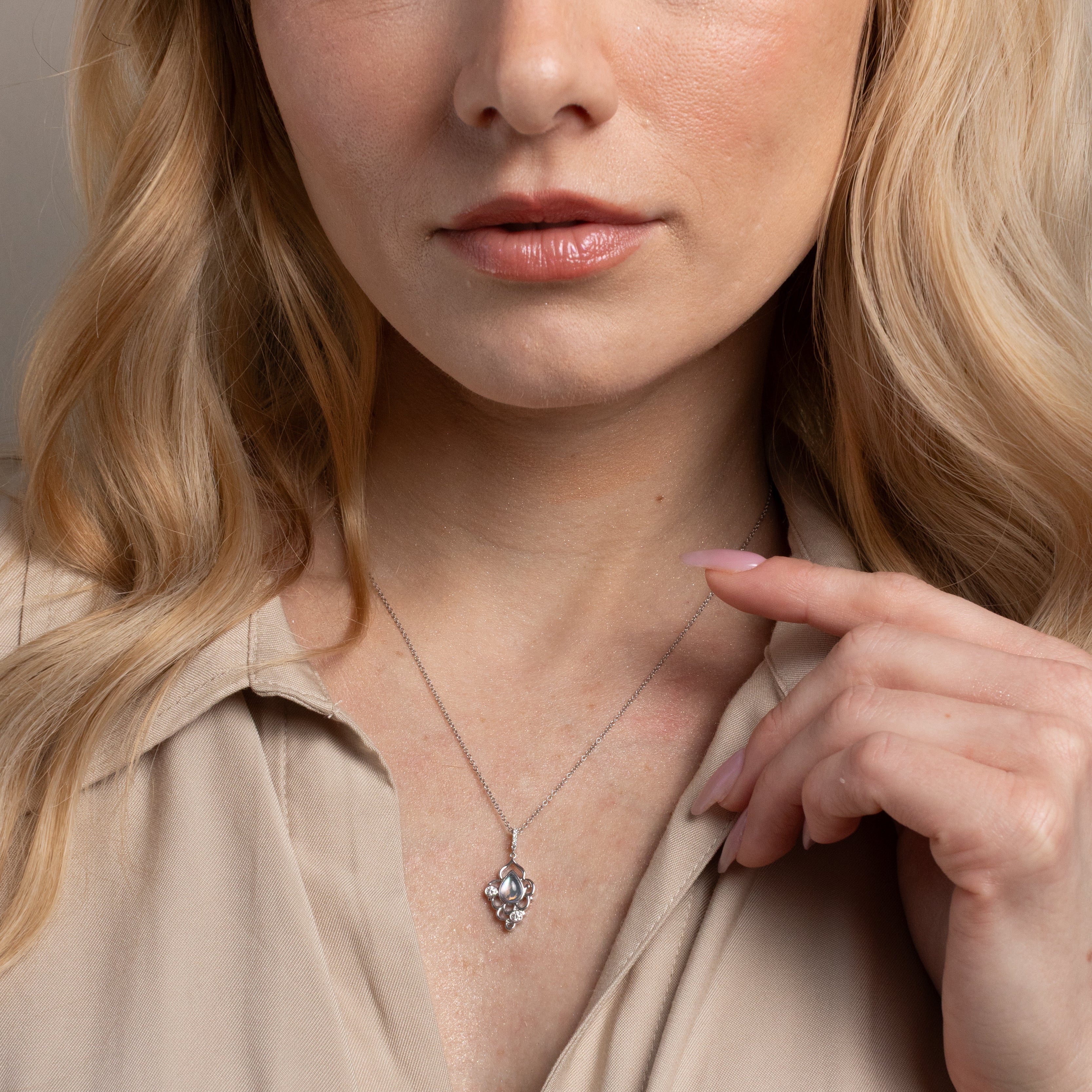 A close-up of a blonde woman wearing a delicate silver pendant necklace and a beige button-up shirt, pointing towards the pendant.