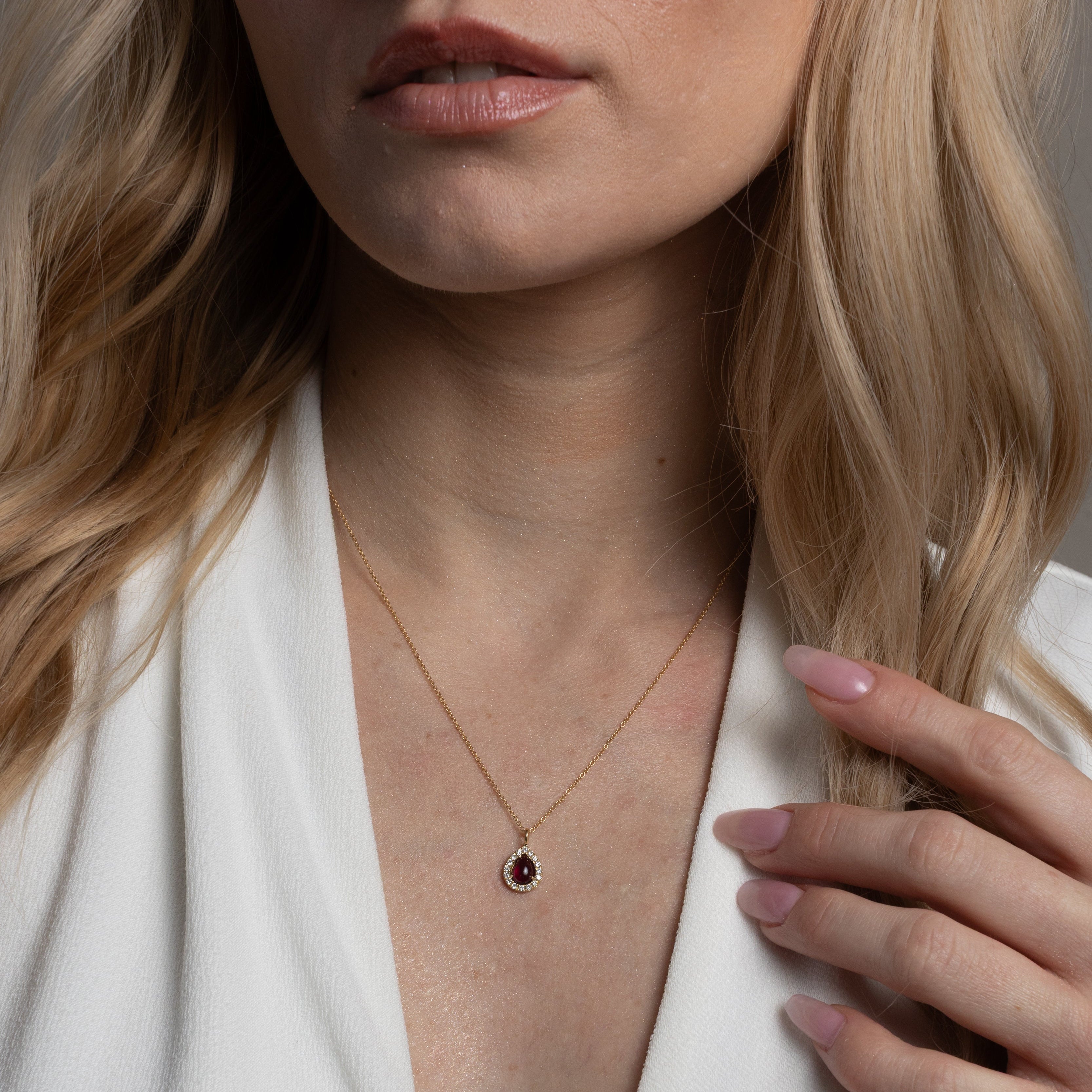 A close-up of a woman wearing a delicate gold necklace with a small dark teardrop-shaped stone pendant, styled with a white V-neck top.