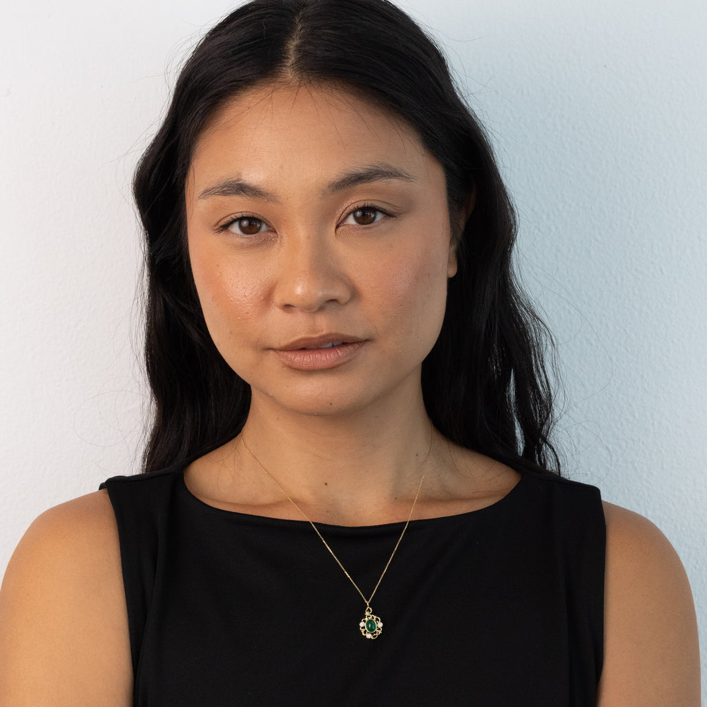 A woman with long dark hair wearing a black sleeveless top and a delicate gold necklace with a green and clear stone pendant, standing against a plain light gray wall.