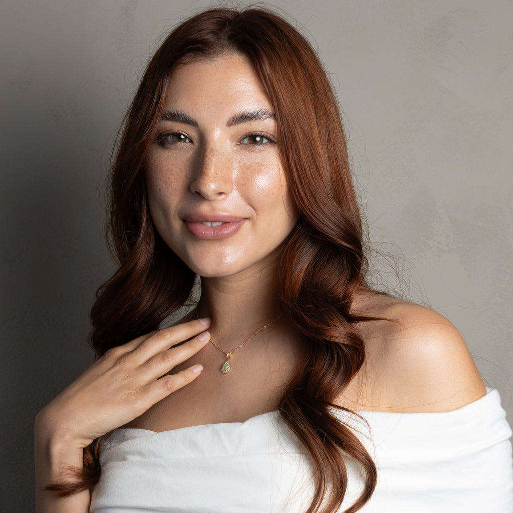 A woman with long wavy brown hair wearing a white off-the-shoulder top and a delicate gold necklace, posing against a neutral gray background.