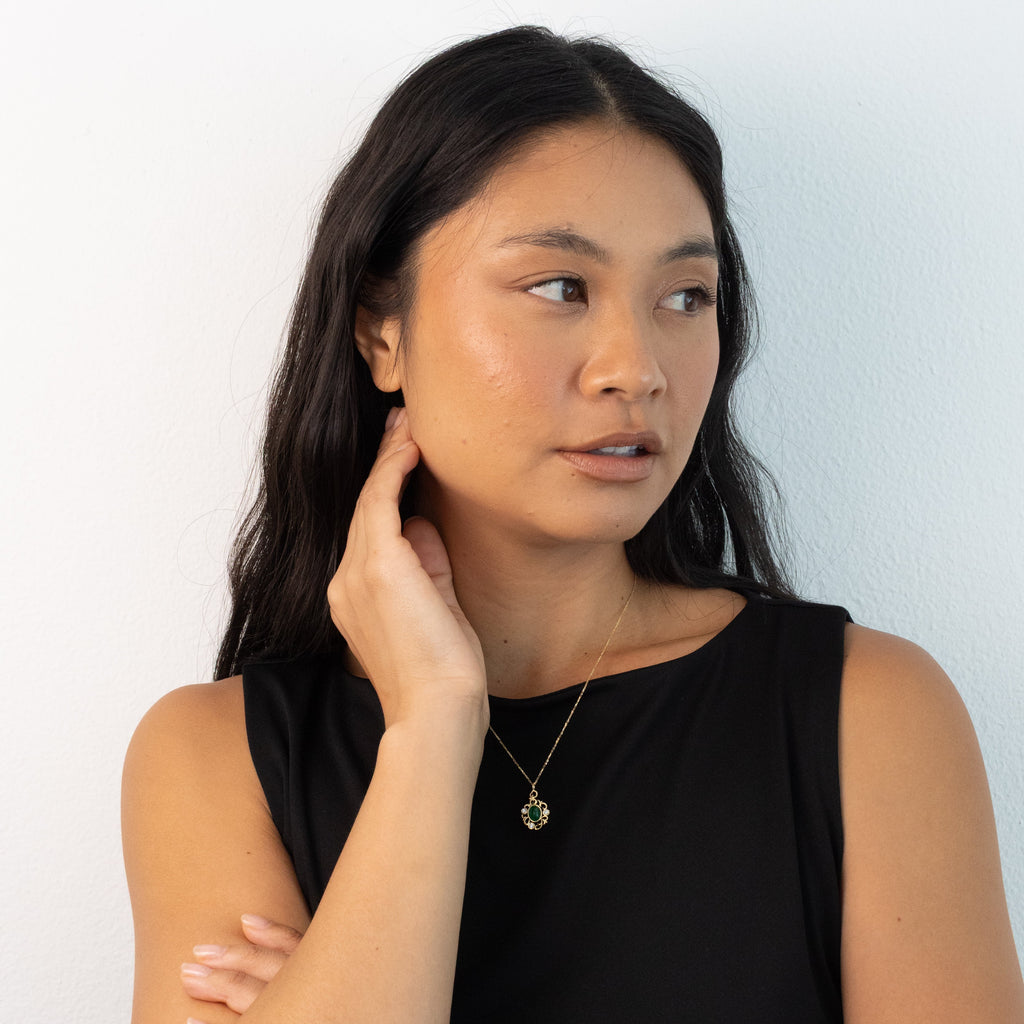 A woman with long dark hair wearing a black sleeveless top and a delicate gold necklace, posing against a plain white wall.