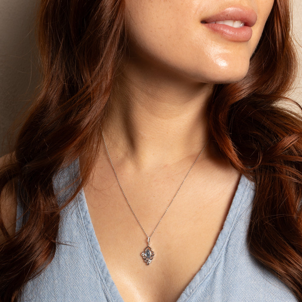 A close-up of a woman wearing a delicate silver pendant necklace with a small floral or starburst design, paired with a light blue denim top.