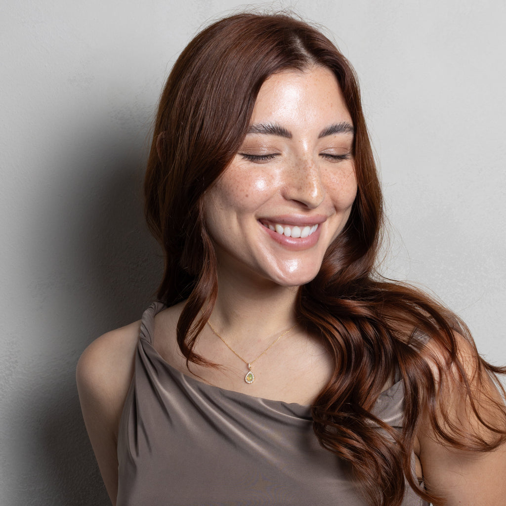 A woman with long brown hair smiling, wearing a taupe-brown ruched one-shoulder top and a delicate gold necklace, posing against a neutral gray wall.