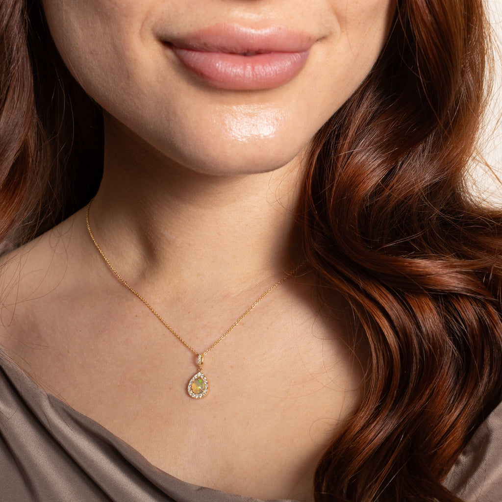 A close-up of a woman wearing a delicate gold chain necklace with a small teardrop-shaped pendant featuring a yellow and green patterned center, paired with a brown satin top.