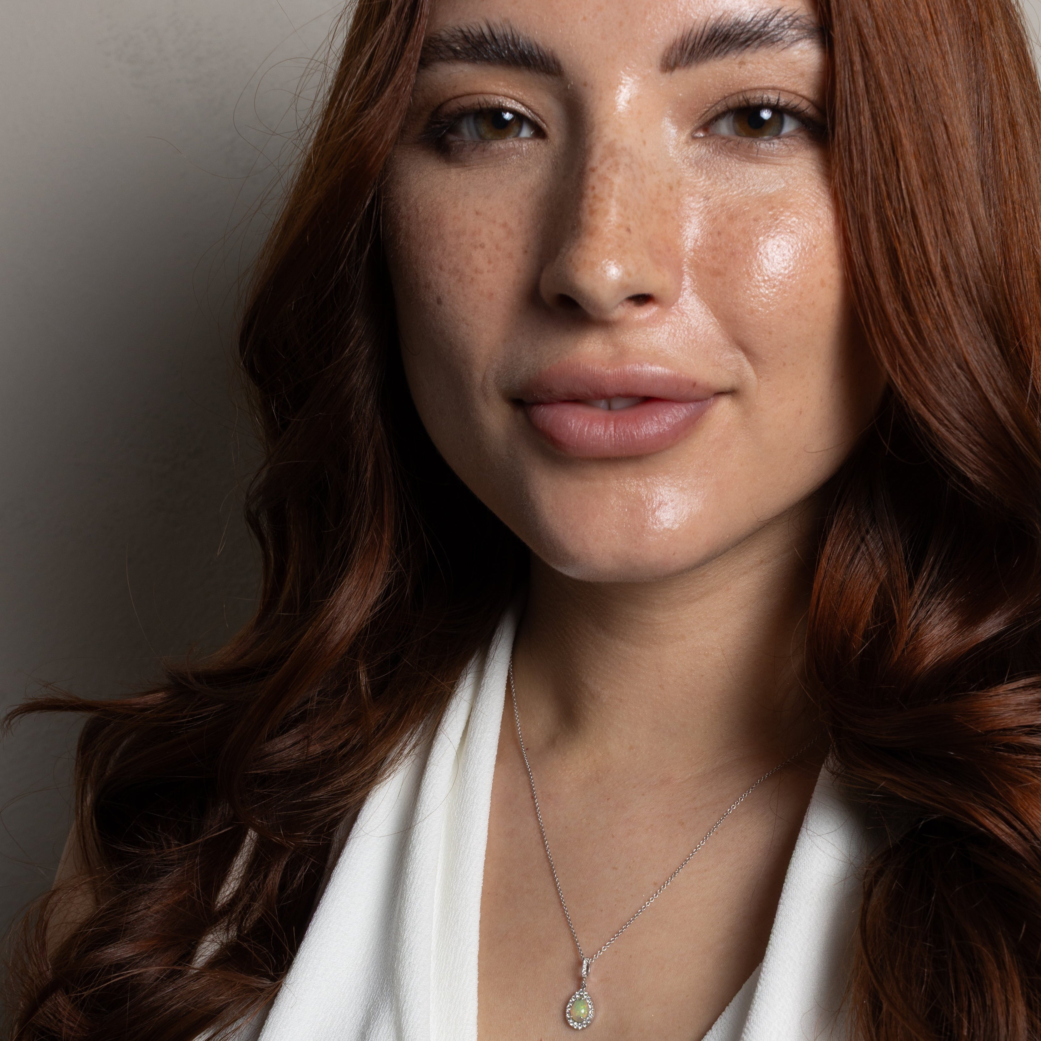 A close-up portrait of a woman with long wavy brown hair wearing a delicate silver necklace with a small oval pendant, paired with a white draped top against a neutral gray background.