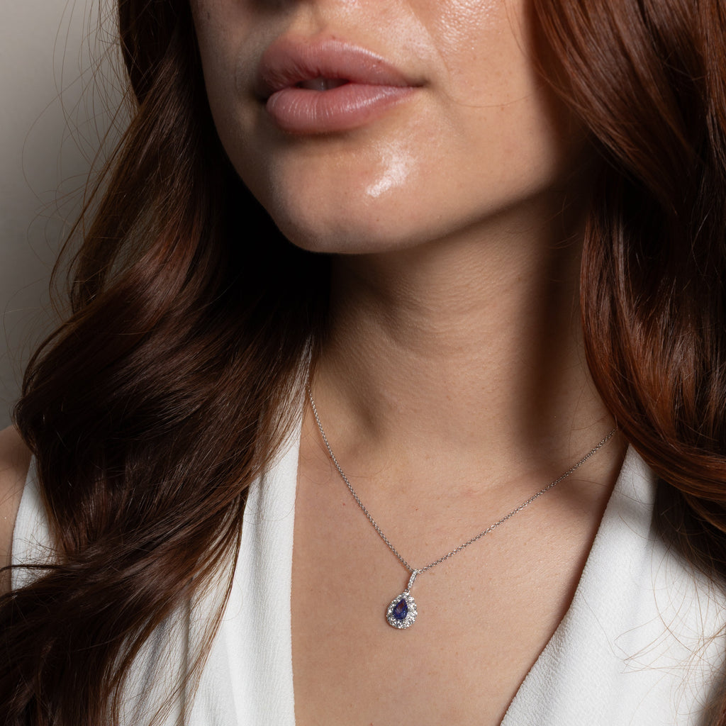 A close-up of a woman wearing a delicate silver chain necklace with a teardrop-shaped blue gemstone pendant, styled with a white V-neck top.