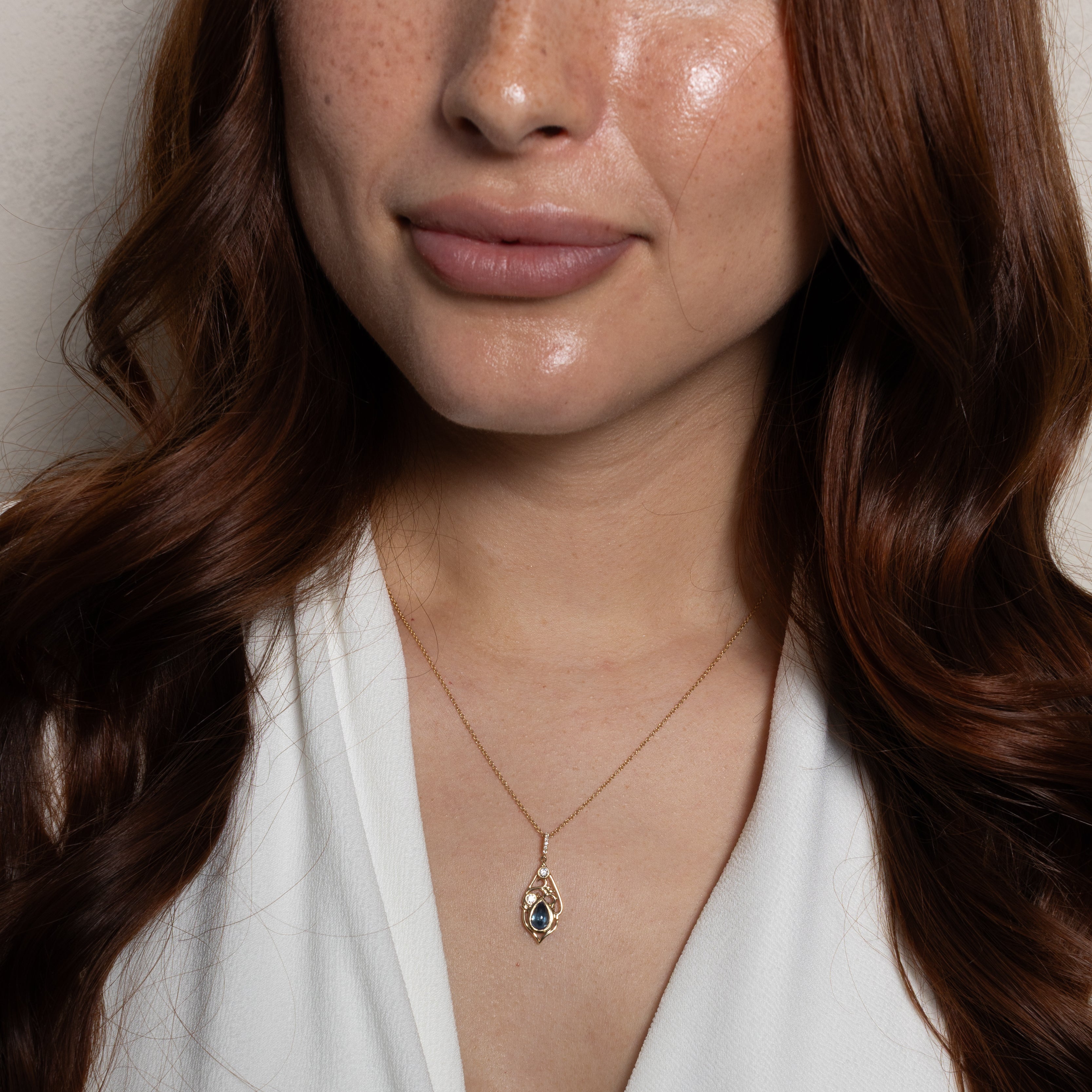 A close-up of a woman with long wavy brown hair wearing a delicate gold chain necklace with a teardrop-shaped pendant featuring a dark blue stone, paired with a white V-neck top.