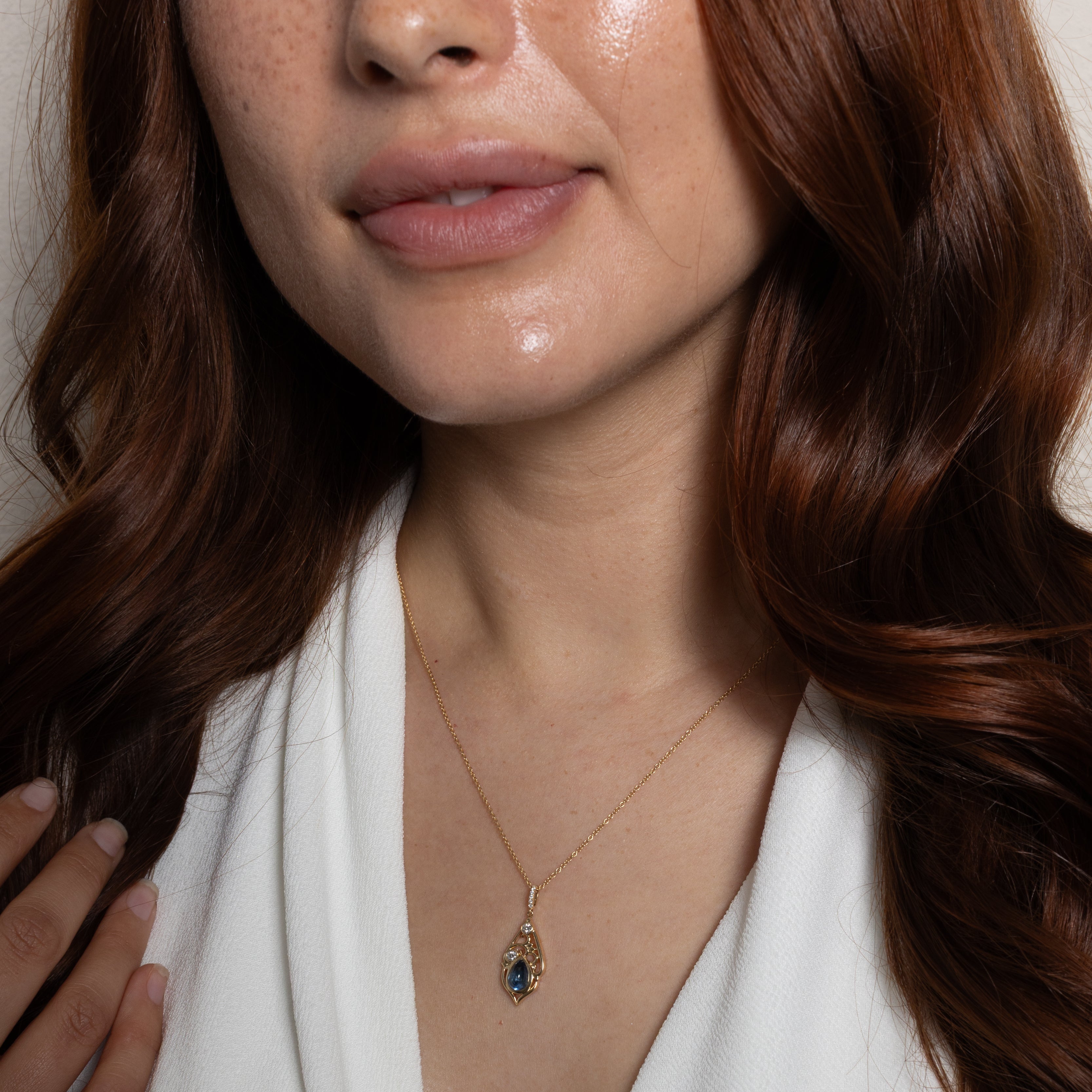 A close-up of a woman wearing a delicate gold necklace with a teardrop-shaped dark blue gemstone pendant, styled with a white V-neck top.