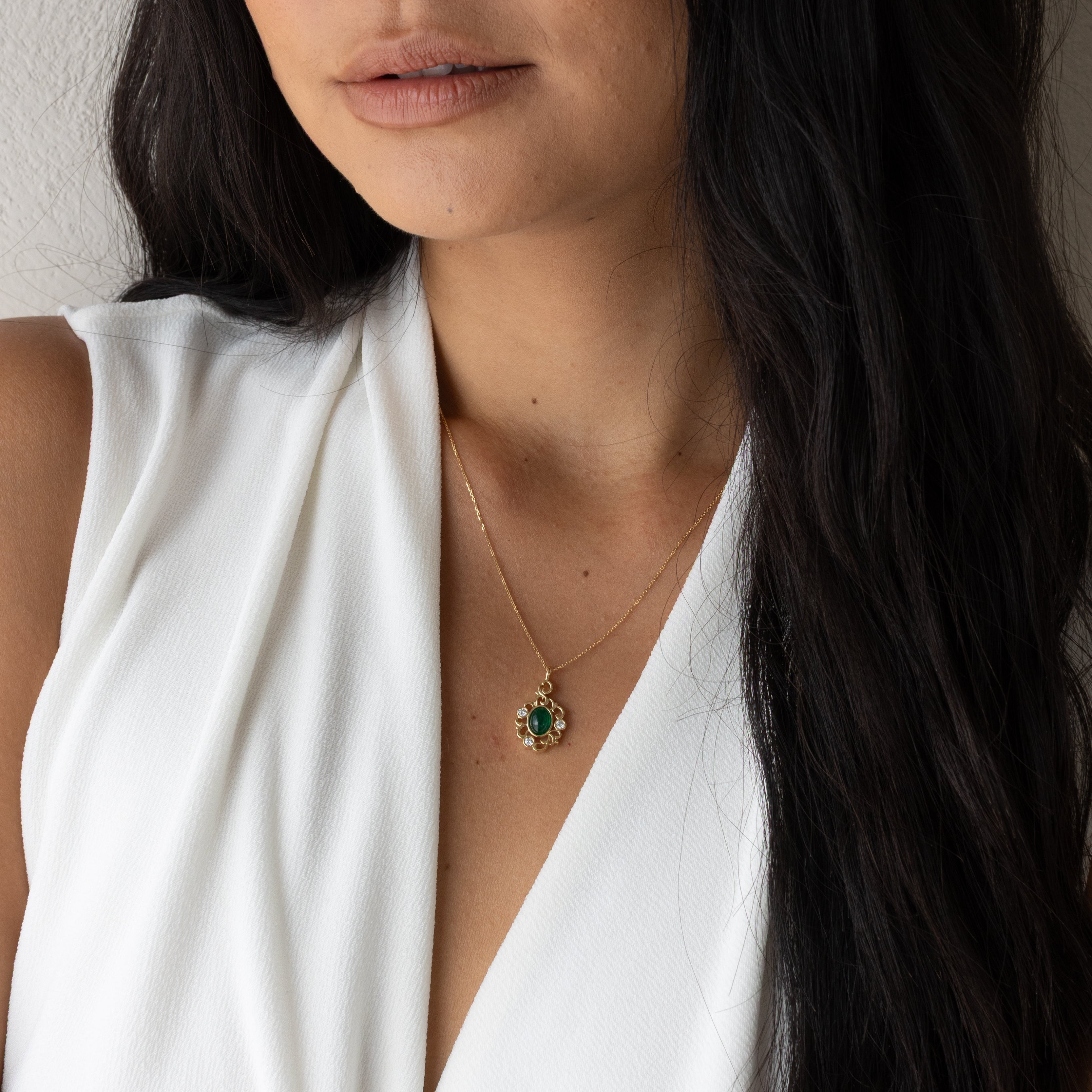 A close-up of a woman with long dark hair wearing a white sleeveless top and a delicate gold necklace with a green and gold pendant, set against a neutral light gray background.