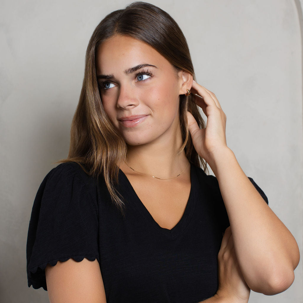 A woman with long brown hair wearing a black V-neck top with scalloped short sleeves, posing against a light gray textured background.