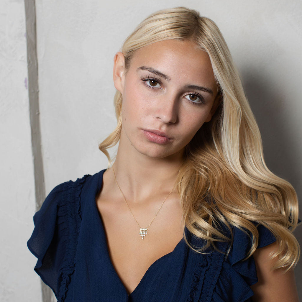 A blonde woman wearing a navy blue ruffled top and a delicate silver necklace, posing against a light gray textured wall.