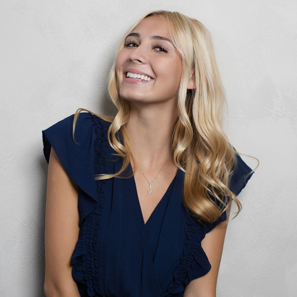 A smiling blonde woman wearing a navy blue ruffled sleeveless top, posing against a light gray textured wall.