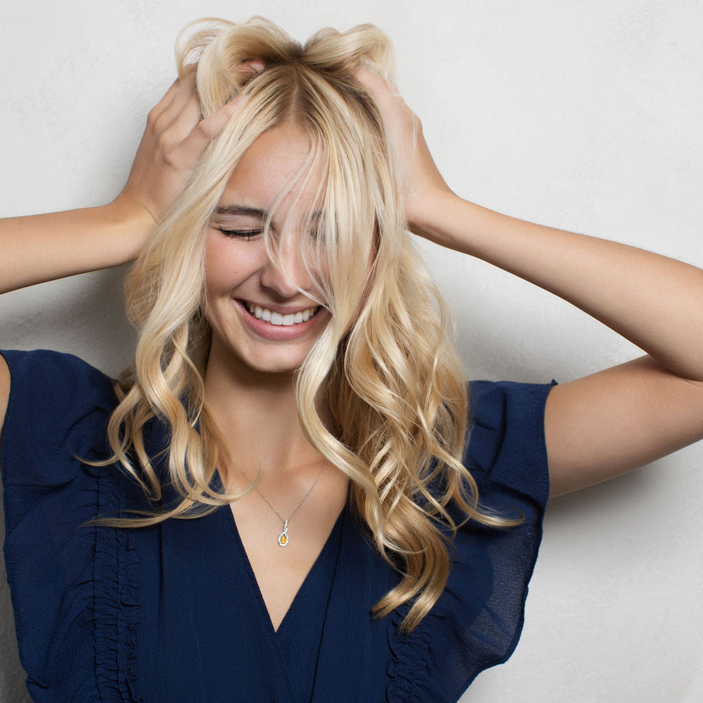 A smiling blonde woman with her hands in her hair, wearing a navy blue ruffled top, posing against a light gray textured wall.