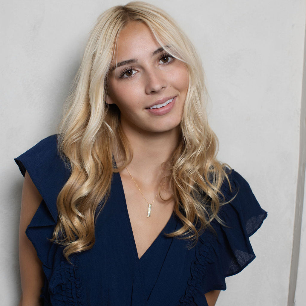 A blonde woman with wavy hair wearing a navy blue ruffled dress and a delicate gold necklace, posing against a light gray textured wall.