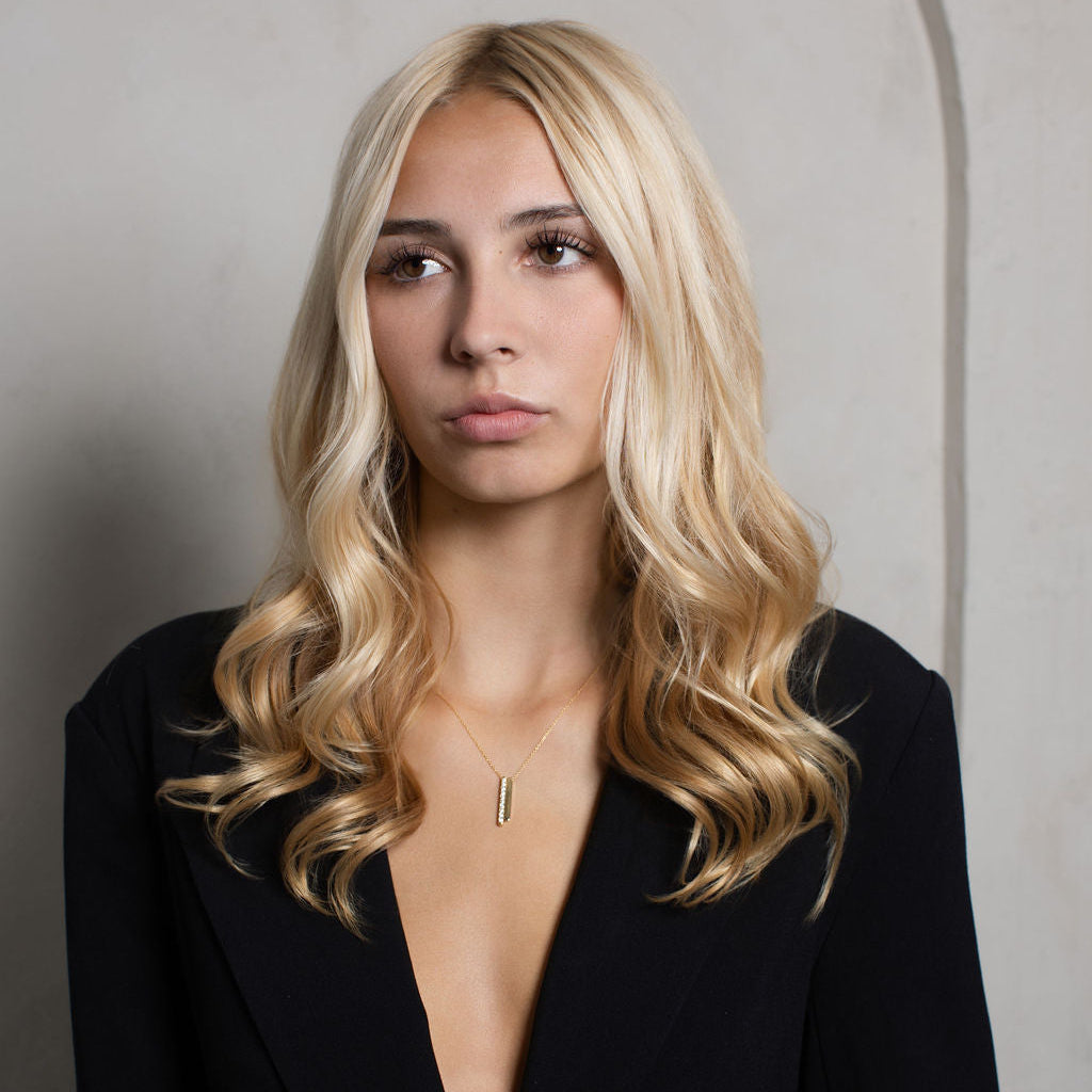 A blonde woman with wavy hair wearing a black deep V-neck blazer and a delicate gold pendant necklace, posing against a textured light gray wall.