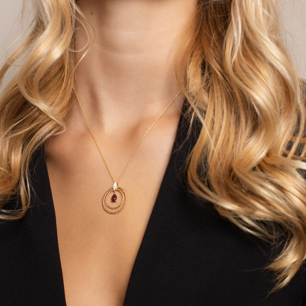 A close-up of a woman wearing a delicate gold necklace with a circular pendant and a small dark red center stone, styled with a black deep V-neck top.