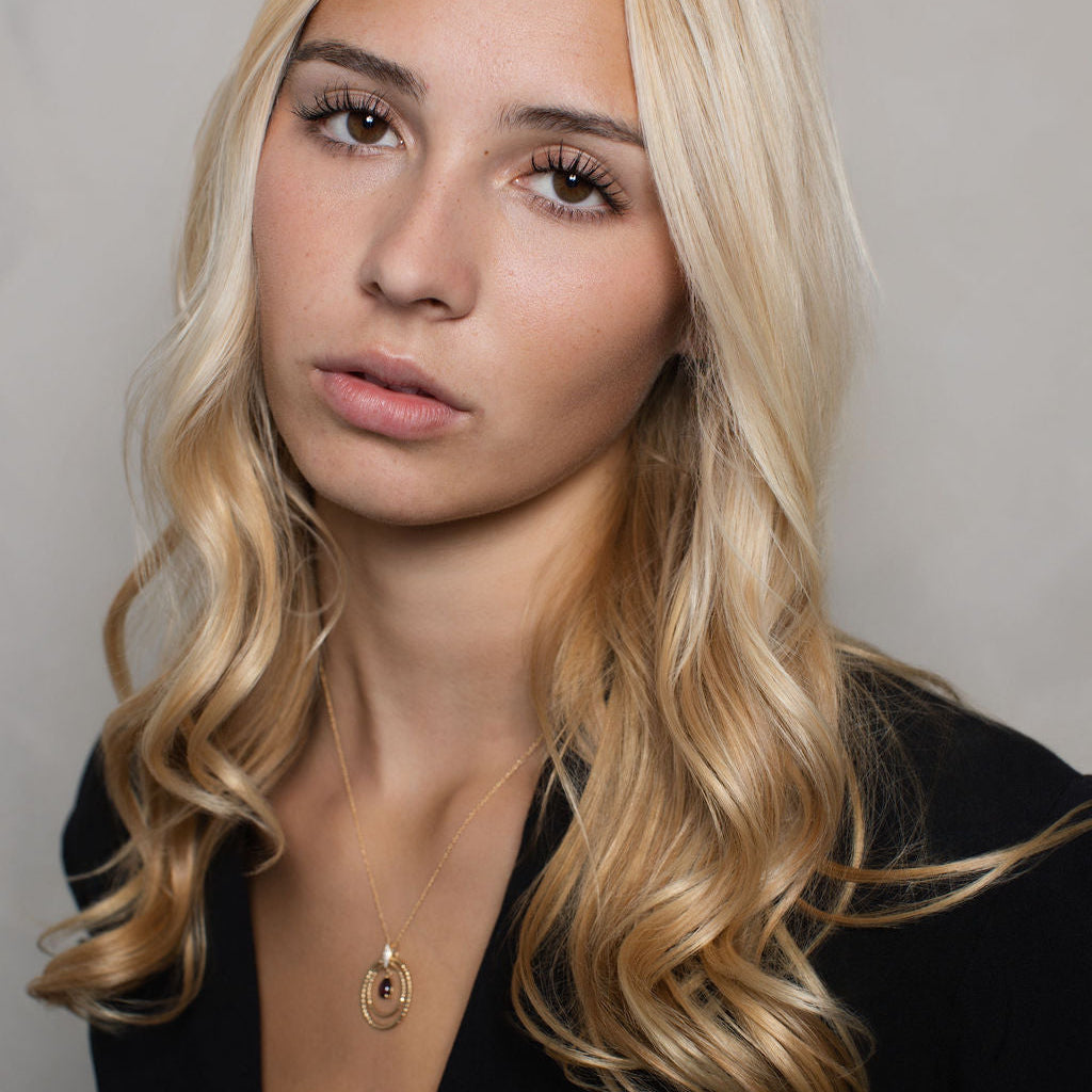 A close-up portrait of a blonde woman with wavy hair, wearing a black top and a delicate gold pendant necklace, set against a plain gray background.