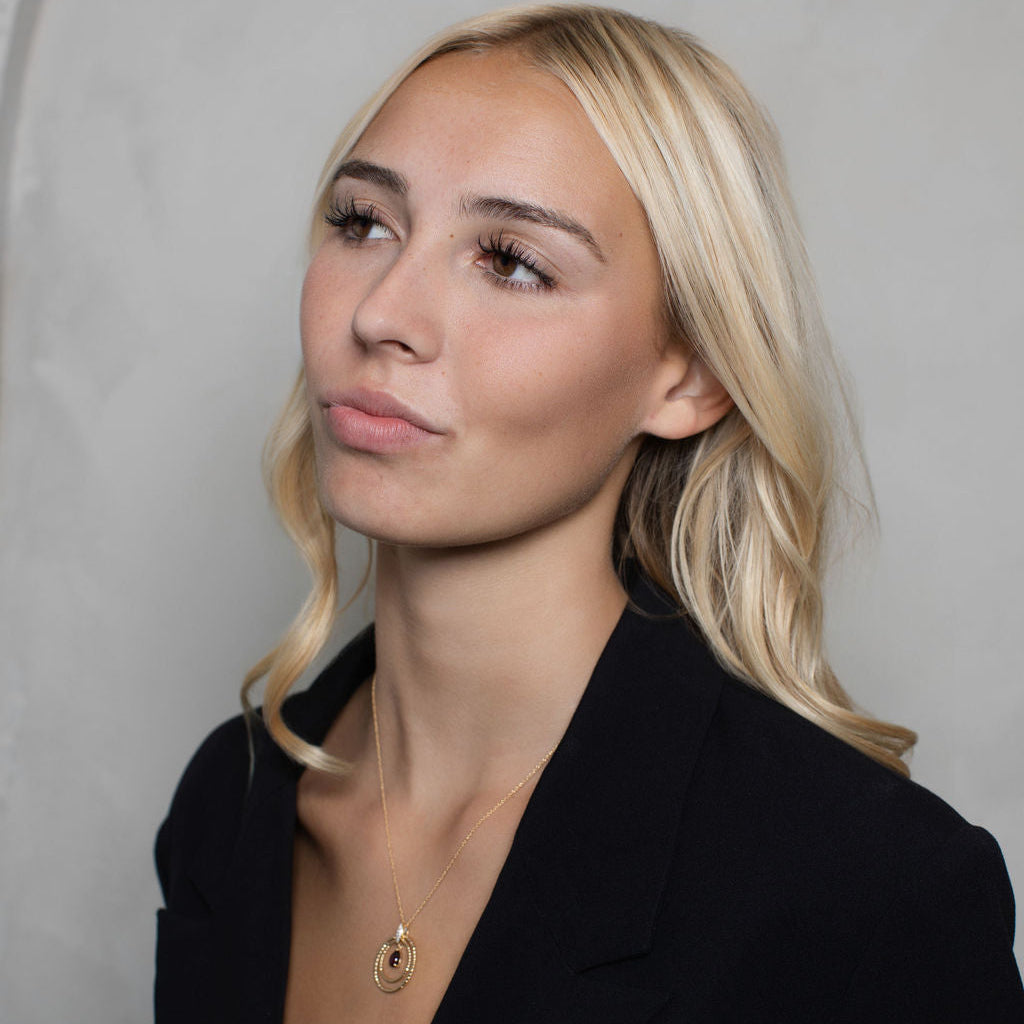 A blonde woman wearing a black blazer and a delicate gold pendant necklace, photographed against a neutral gray background.