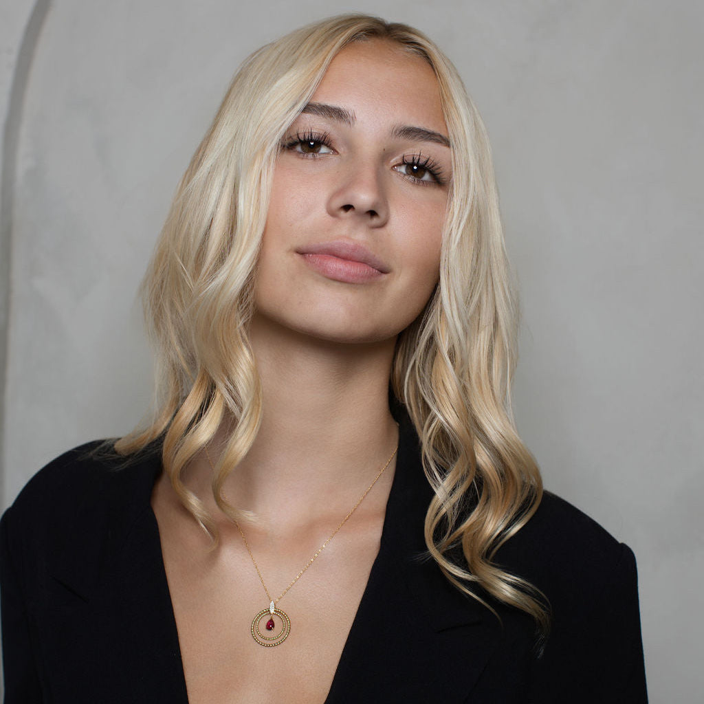 A blonde woman with wavy hair wearing a black blazer and a delicate gold pendant necklace with a red stone, posing against a textured gray studio background.
