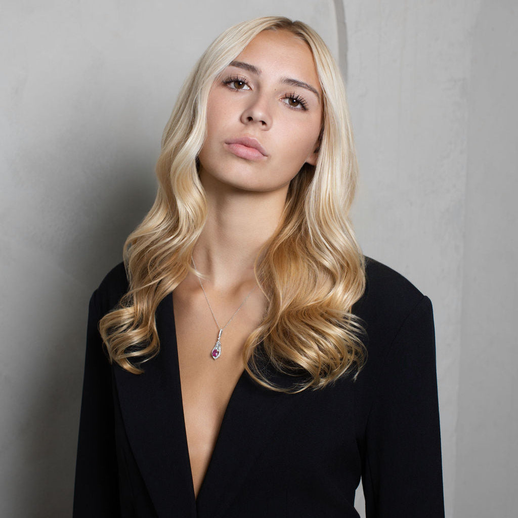 A blonde woman with wavy hair wearing a black blazer and a delicate silver necklace with a purple gemstone, posing against a textured gray wall with a subtle arch design.