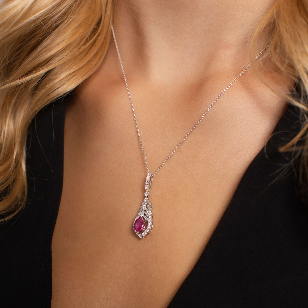A close-up of a model wearing a silver teardrop pendant necklace with a central pink gemstone and small clear accent stones, paired with a black top.