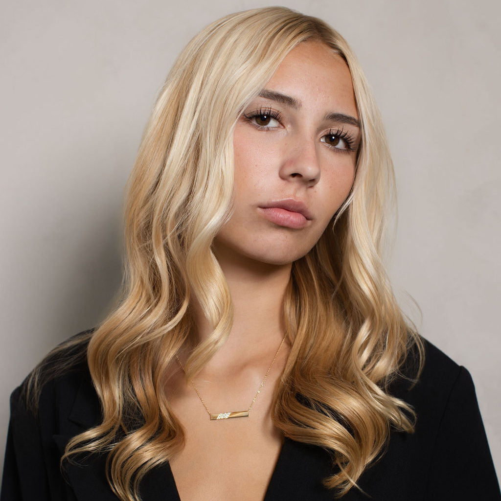 A blonde woman with wavy hair wearing a black blazer and a delicate gold bar necklace, photographed against a neutral gray background.
