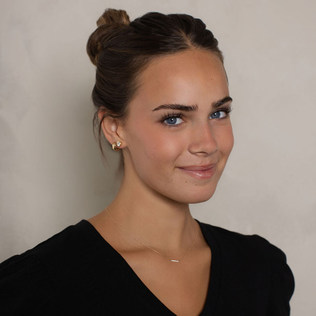 A woman with her hair in a bun wearing a black V-neck top, shown from the chest up against a light gray textured background.