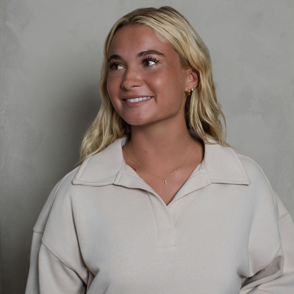 A smiling blonde woman wearing a light beige long-sleeve polo sweatshirt, standing against a textured gray wall.