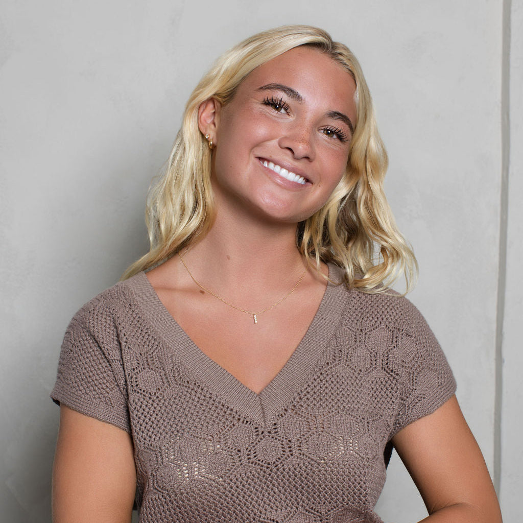A smiling blonde woman wearing a brown short-sleeve knit V-neck top, standing against a light gray textured wall.