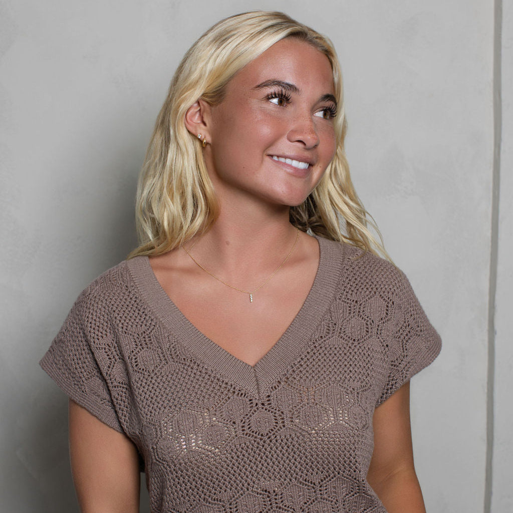 A smiling blonde woman wearing a taupe brown open-knit V-neck short-sleeve top, standing against a textured light gray wall.