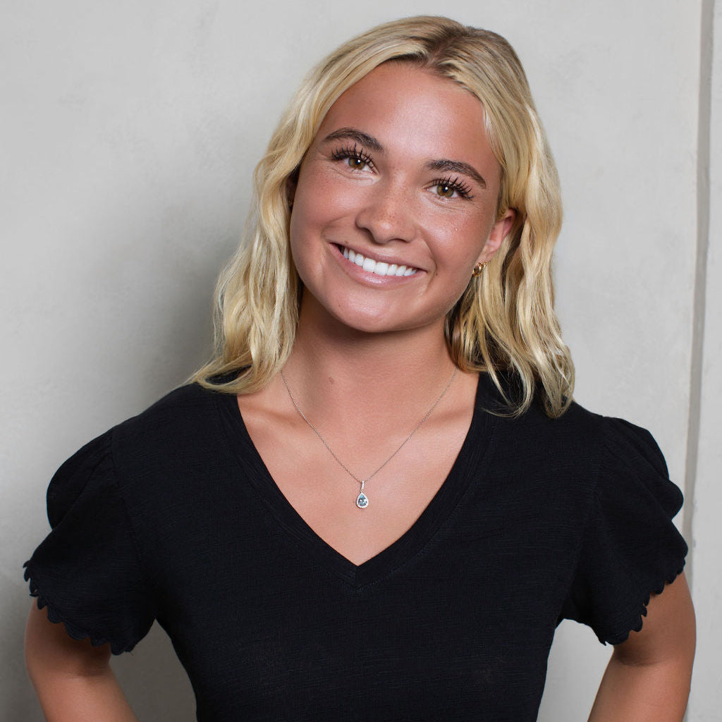 A smiling blonde woman wearing a black short-sleeve V-neck top with ruffled sleeves, standing against a light gray wall.