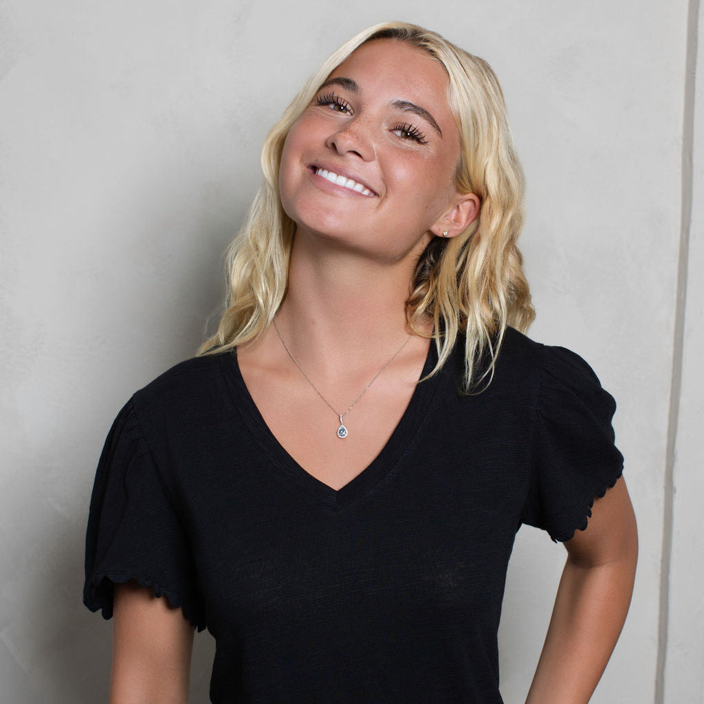 A smiling blonde woman wearing a black short-sleeve V-neck top with ruffled edges, standing against a light gray textured wall.